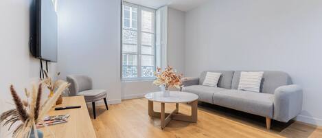 Un moderno salón con un sofá gris, una mesa redonda de centro, un televisor montado en la pared y una ventana con vistas al exterior.