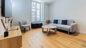 A modern living room with a grey sofa, a round coffee table, a grey armchair, and a wooden side table with a remote control and magazines. - Luxury 3-bedroom apartment in hyper centre, air con, lift (Bordeaux)