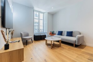 A modern living room with a grey sofa, a round coffee table, a grey armchair, and a wooden side table with a remote control and magazines. - Luxury 3-bedroom apartment in hyper centre, air con, lift (Bordeaux)