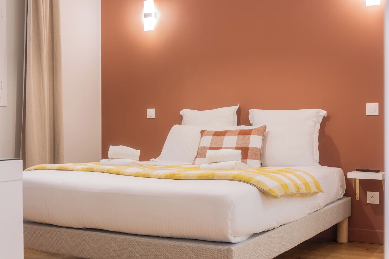 Una cama con ropa de cama blanca y una manta a rayas amarillas y blancas, un almohadón blanco y un almohadón de cuadros marrones y blancos.