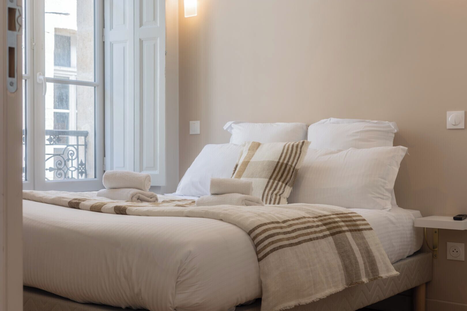 A neatly made bed with white linens and striped blankets, a bedside table with a phone, and a window with a view of buildings.