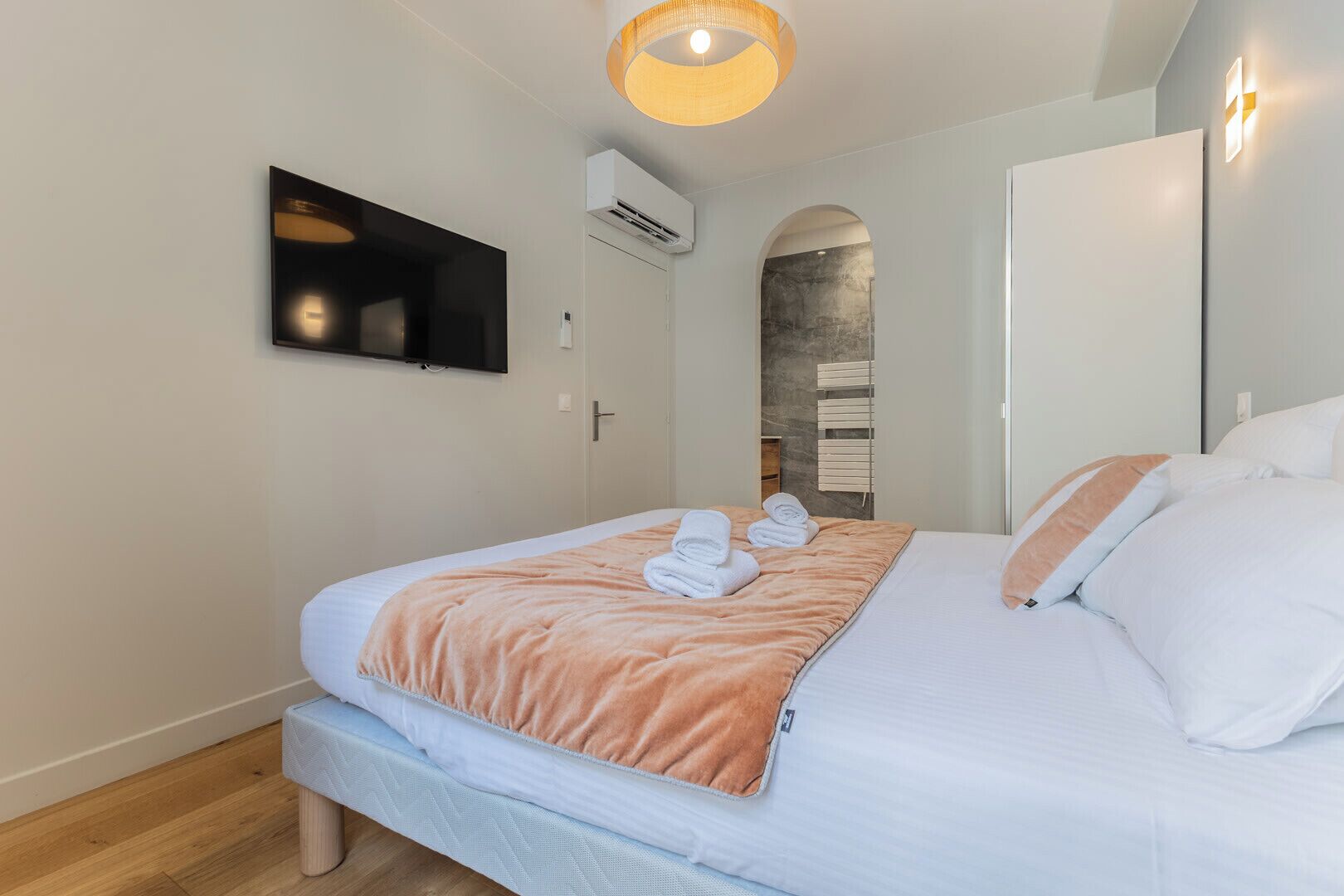 A modern bedroom with a bed, a flat-screen TV, an air conditioning unit, and a wall-mounted light fixture.
