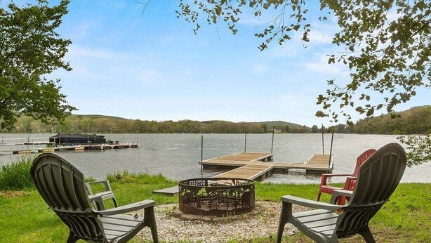 Blick auf einen See mit zwei Adirondack-Stühlen, einer Feuerstelle und einem Steg, der ins Wasser führt.