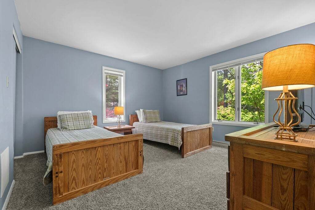 A bedroom with two wooden beds, a desk, and two windows with views of greenery.