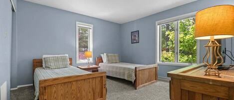 A bedroom with two wooden beds, a desk, and two windows with views of greenery.