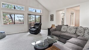 Ein geräumiges Wohnzimmer mit einem grauen Ecksofa, einem Glastisch, einem Ledersessel und großen Fenstern mit Blick auf Bäume und ein Gebäude.