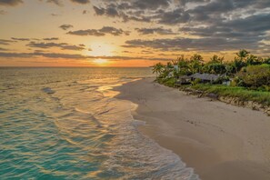 Une plage au coucher du soleil, avec une mer calme et une rangée de maisons offrant une vue sur l’océan.