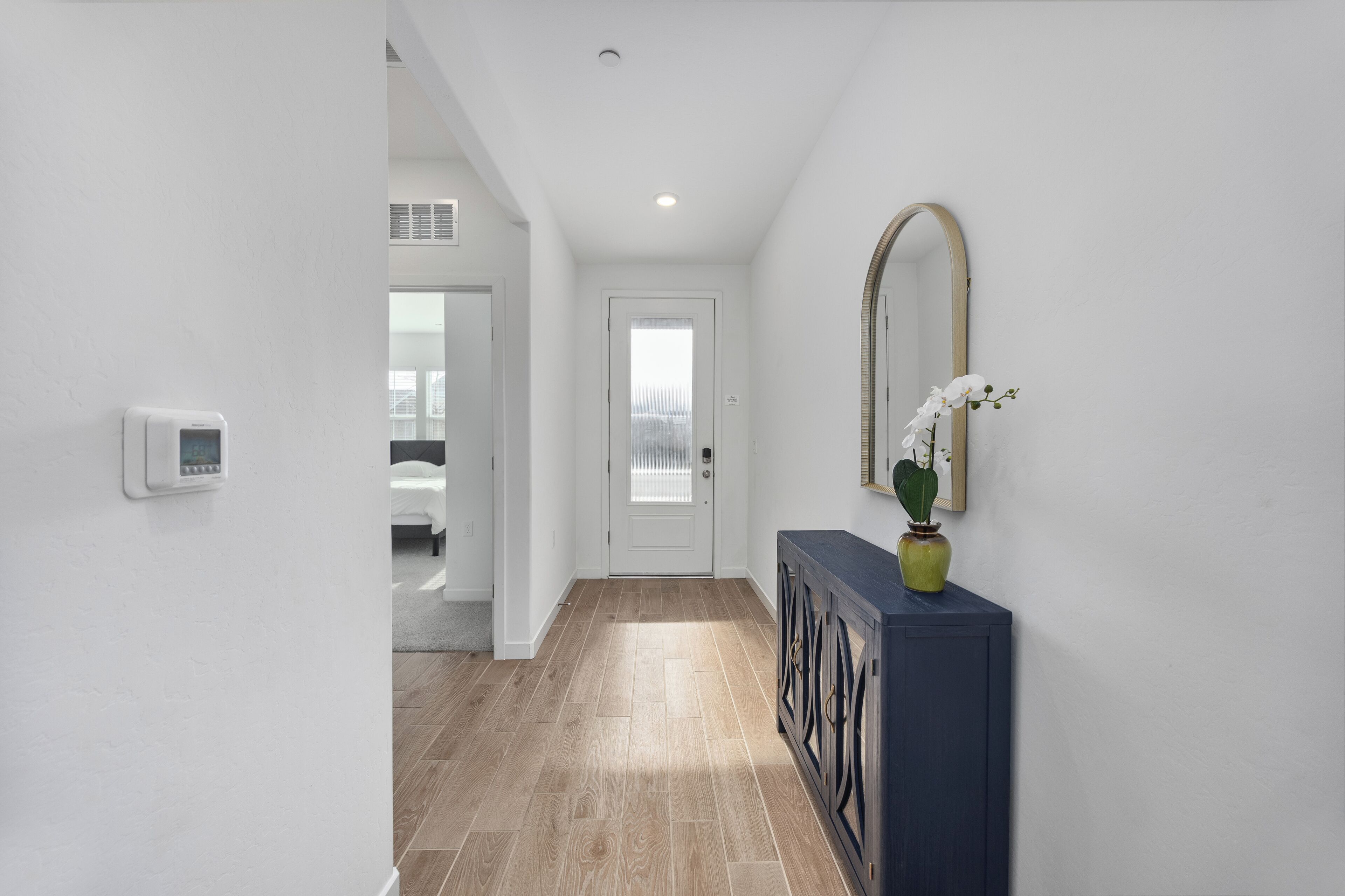 A hallway with a white door, a thermostat, a console table with a vase, and a mirror on the wall.