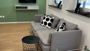 Un salon moderne avec un canapé gris, un coussin à motifs noir et blanc, une petite table d’appoint noire et une étagère métallique verte.