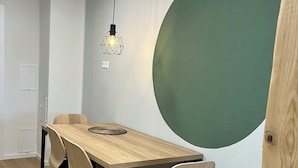 Un espace repas comprenant une table en bois, quatre chaises, un luminaire suspendu et une grande fresque murale verte.