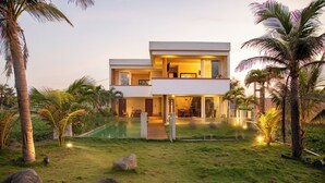 A modern two-story house with a swimming pool, surrounded by palm trees and rocks.