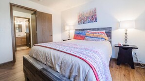 Ein Schlafzimmer mit einem Bett, zwei Nachttischen, einer Sitzbank, einer Tür und einem Gemälde an der Wand.