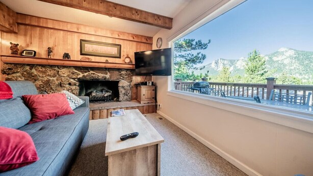 Ein gemütliches Wohnzimmer mit Kamin aus Stein, einem grauen Sofa mit roten Kissen, einem Holztisch und einem großen Fenster mit Blick auf eine Terrasse und Berge.