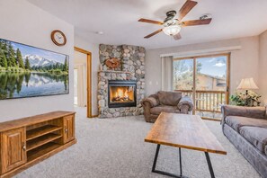 A living room with a stone fireplace, a ceiling fan, a large window, and a wooden coffee table.