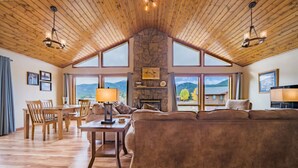 Ein geräumiges Wohnzimmer mit Kamin aus Stein, Holzboden und großen Fenstern mit Blick auf die Berge.