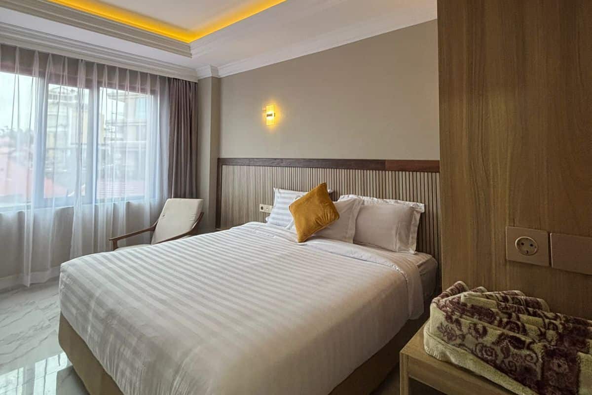 A hotel room with a large bed, a bedside table, a lamp, and a view of the outside through a window with curtains.