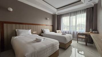 A hotel room with two beds, a wooden headboard, a desk, and a large window with curtains.