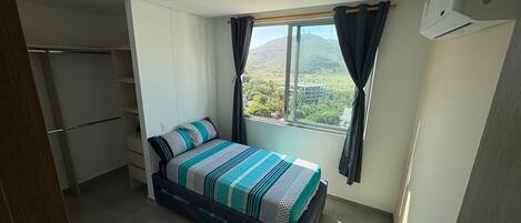 Quarto com cama, guarda-roupa, janela com cortinas e ar condicionado.