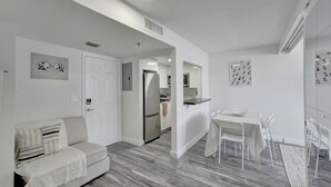 Uma sala de estar moderna, com decoração em branco, incluindo um sofá, uma área de refeições com mesa e cadeiras, e uma cozinha ao fundo.
