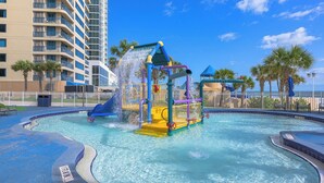 A water park with a water slide and splash pad, surrounded by palm trees and a building. - Palm Royale - Ocean Walk (Daytona Beach)
