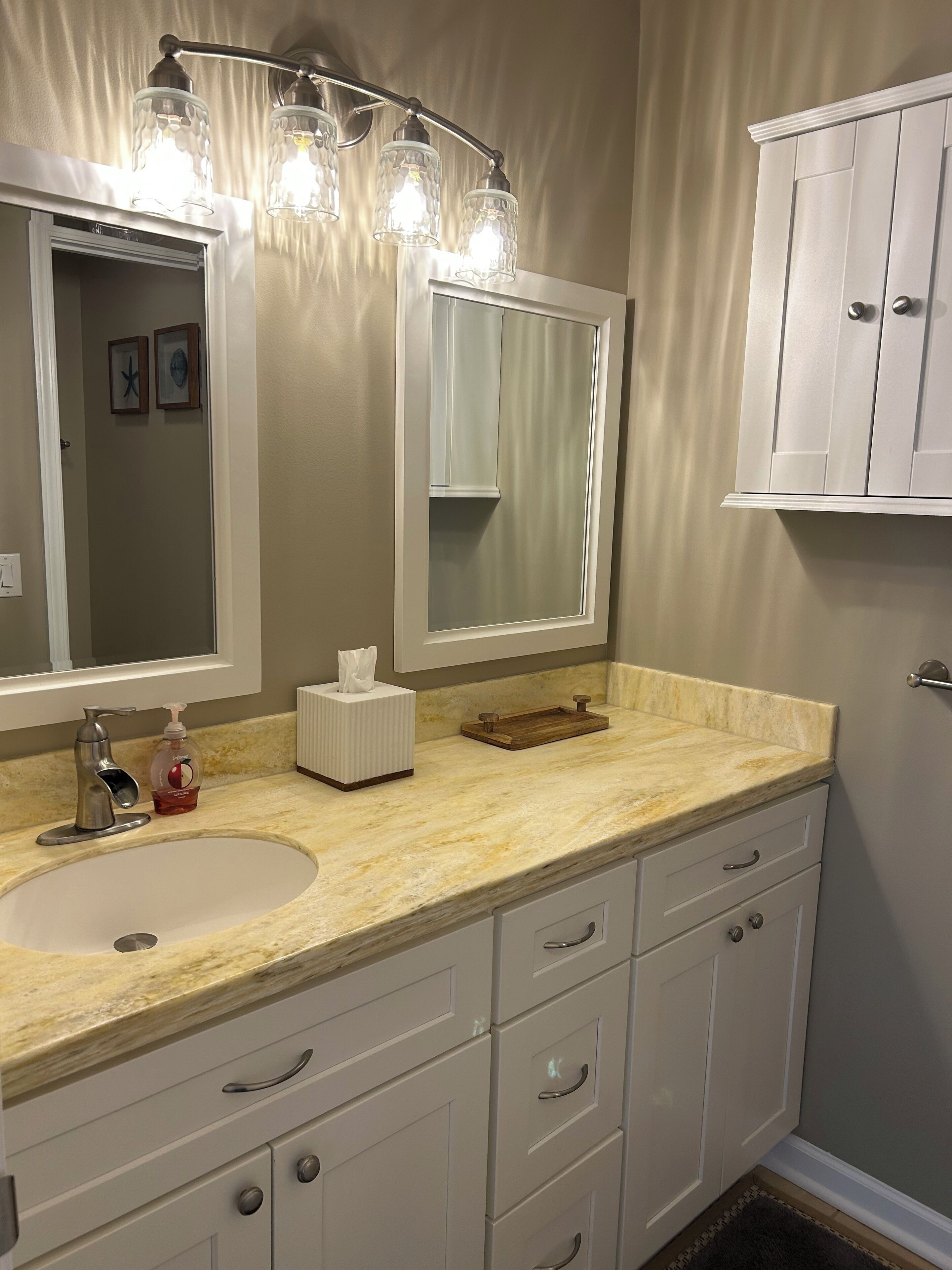 A bathroom with a white sink, vanity, and cabinet. There is a mirror with lights above it, and a towel rack on the wall.