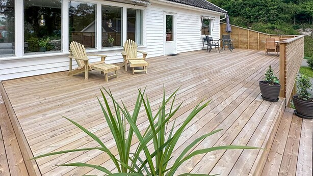 Una terraza de madera con sillas y una mesa, rodeada por una valla de madera y plantas en macetas.