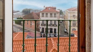 Vista a través de una ventana con barandilla de balcón, una calle con coches estacionados y edificios con tejados de tejas rojas.