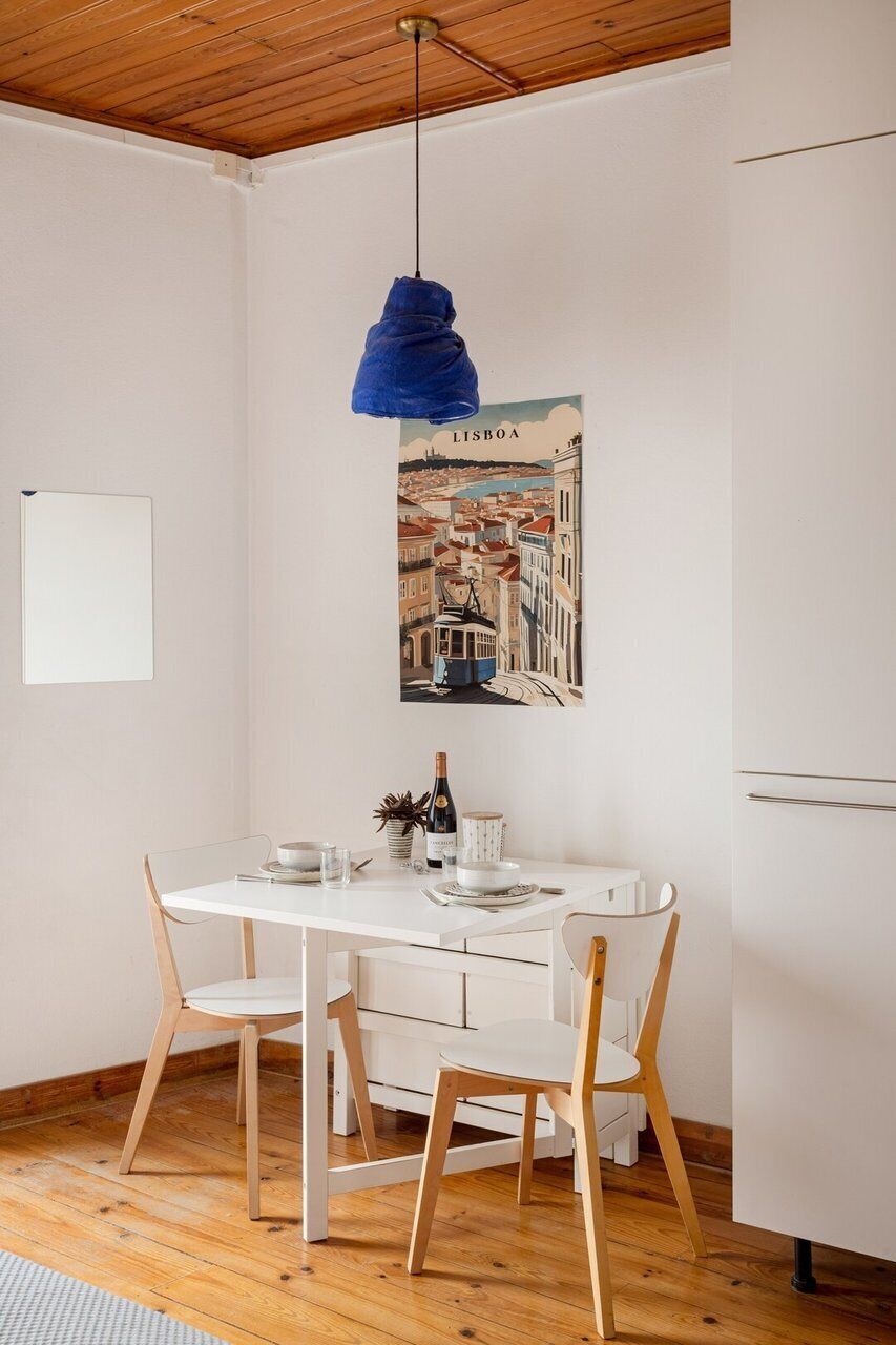 Ein Essbereich mit einem weißen Tisch, zwei Holzstühlen, einer blauen Pendelleuchte, einem an der Wand hängenden Bild von Lissabon und einer Whiteboard-Tafel.