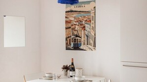 Un comedor con una mesa blanca, dos sillas de madera, una lámpara colgante azul, un cuadro de Lisboa en la pared y una pizarra.