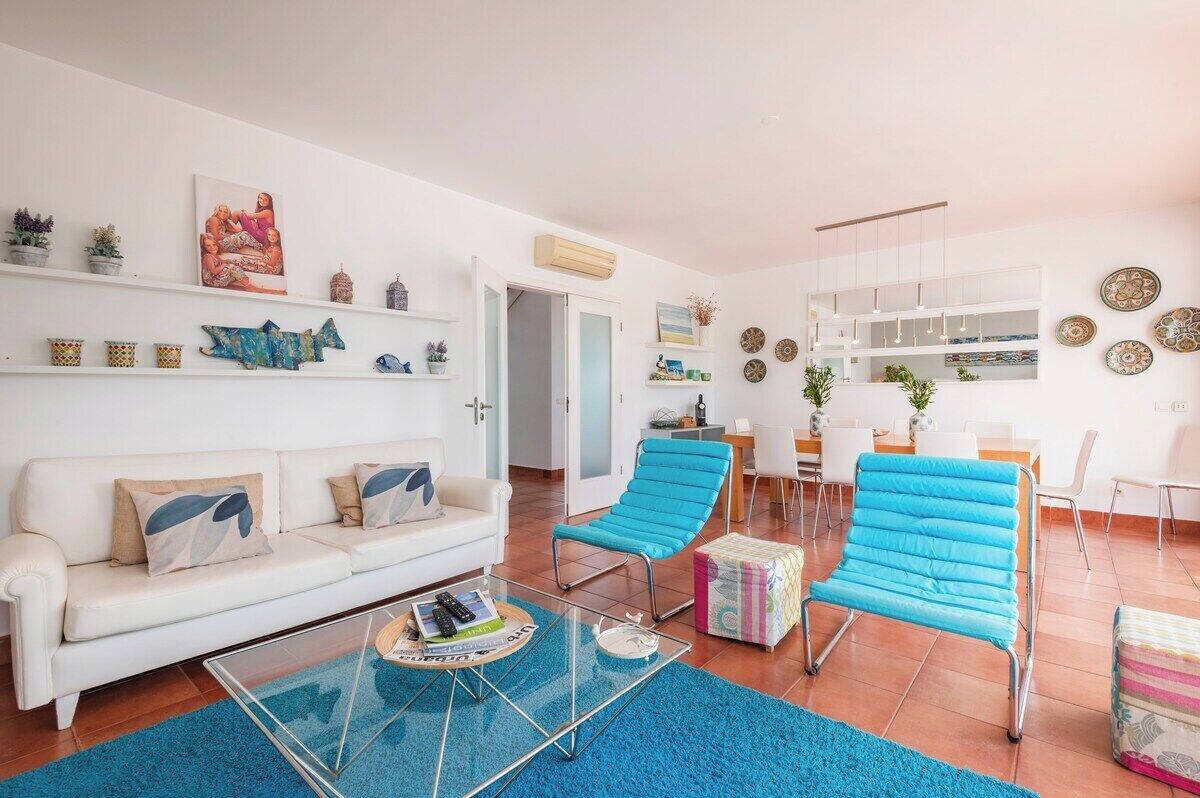 Un salon moderne avec un canapé blanc, des fauteuils bleus, une table basse en verre et, au fond, un coin repas.