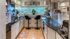A modern kitchen with two black bar stools, a window with a view of greenery, and stainless steel appliances.