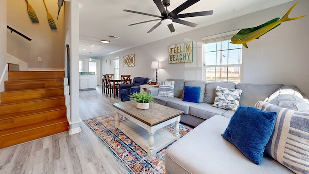 A spacious living room with a wooden staircase, a ceiling fan, a sofa set, and a large window. - Feelin Beachy, Shared Pool, Walk to the Beach (Port Aransas)