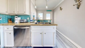 A kitchen with white cabinets, a stainless steel dishwasher, a granite countertop, and a window with a view. - Feelin Beachy, Shared Pool, Walk to the Beach (Port Aransas)