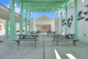 Un espace extérieur abrité, doté de tables de pique-nique et de guirlandes lumineuses, avec un bâtiment blanc en arrière-plan et des plantes en pot disposées le long des murs.