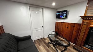 A living room with a black sofa, a glass table, a TV mounted on the wall, and a wooden-paneled wall. - Prime Location 3Bedrooms, 2 Bath,Basement Apt with free parking-Close to Transit (Mississauga)