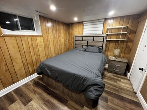 Ein Schlafzimmer mit einer holzverkleideten Wand, einem Bett mit grauer Bettwäsche, einem Nachttisch mit einer Lampe und einem Fenster mit Jalousien.