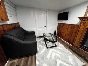 Una sala de estar con un sofá negro, una mesa de centro de vidrio, un tapete estampado, piso de madera, una puerta blanca y un televisor montado en la pared.