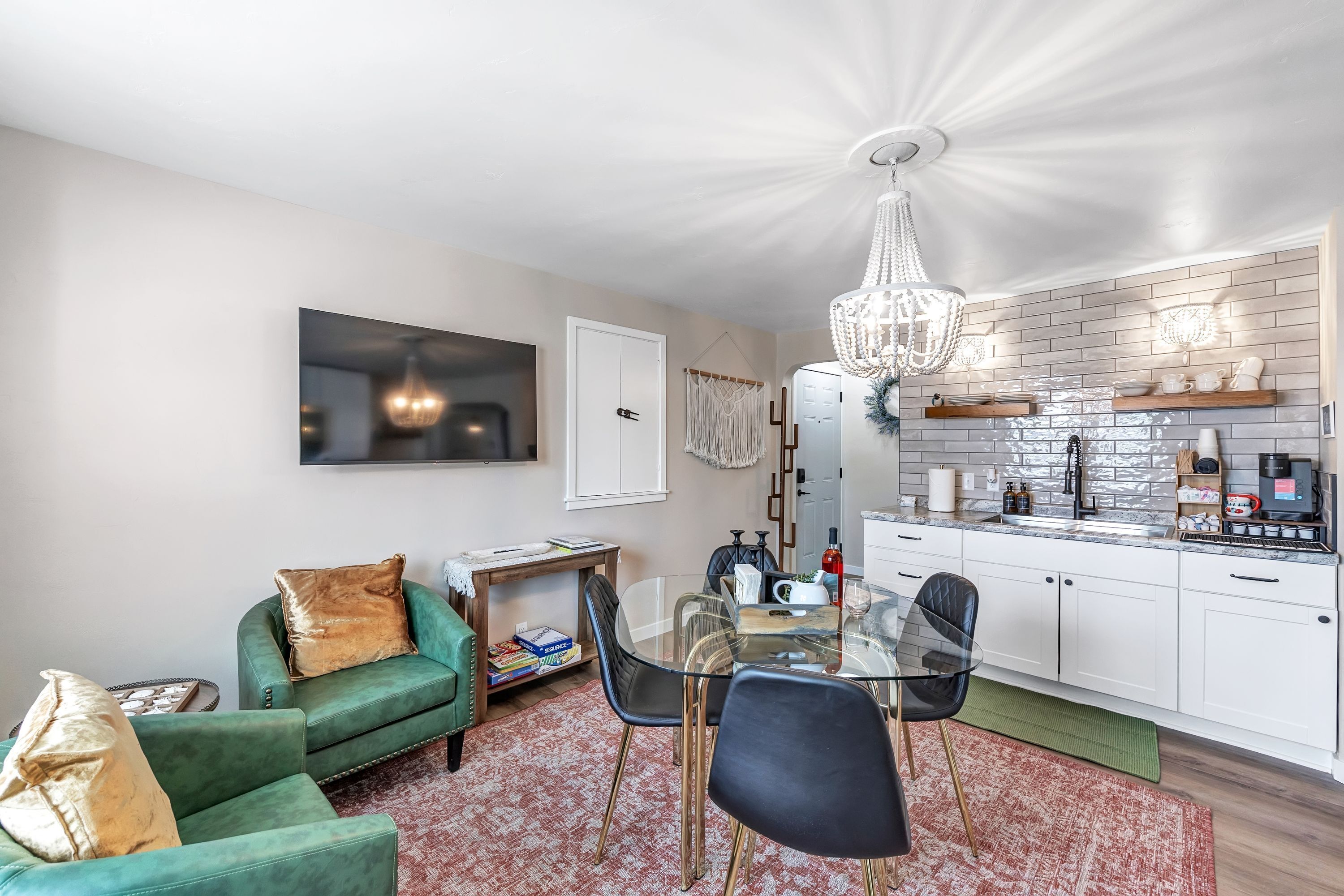 Een moderne woonkamer met een glazen eettafel, een groene bank en een keuken met witte kasten en een metselwerk achterwand.