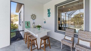 Une cuisine moderne avec un îlot central blanc, des tabourets de bar, des chaises en osier et une grande fenêtre donnant sur l’extérieur.
