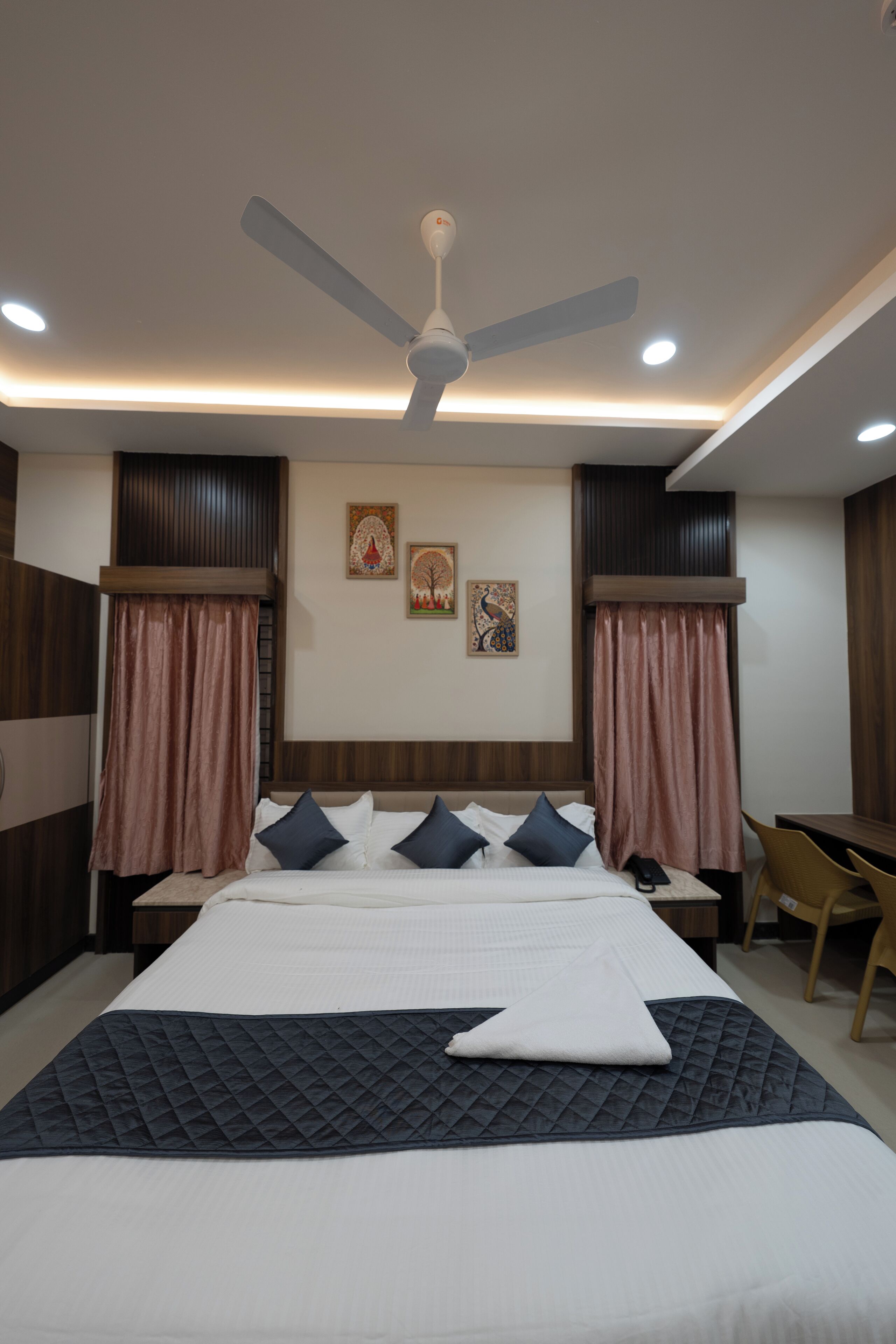 A hotel room with a bed, a desk, a chair, a ceiling fan, and artwork on the wall.