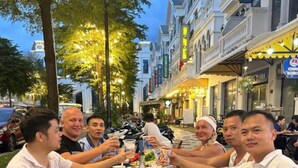 Un groupe de personnes est assis à une table à l’extérieur, devant de la nourriture et des boissons, dans une rue bordée d’immeubles et d’arbres.