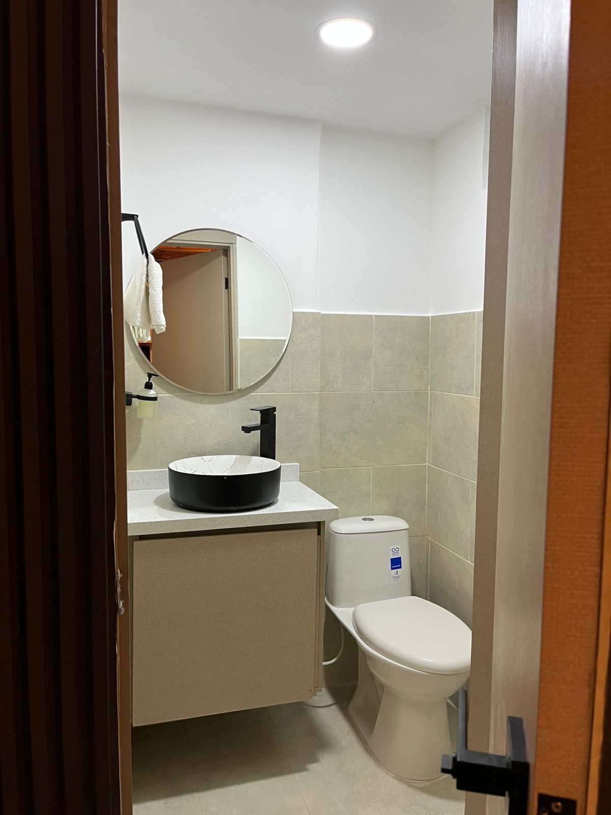 Une petite salle de bain comprenant un miroir rond, un lavabo, des toilettes et un porte-serviettes.