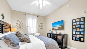 Une chambre à coucher comprenant un lit, une table de chevet, une commode, un ventilateur de plafond et un mur orné d’un tableau encadré et d’un photomontage.