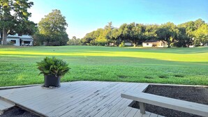 Un campo cubierto de césped con una terraza de madera y un banco, rodeado de árboles y casas.