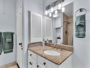 Un baño con lavamanos de encimera de granito, armarios blancos, un espejo grande con iluminación y un perchero con toallas verdes.