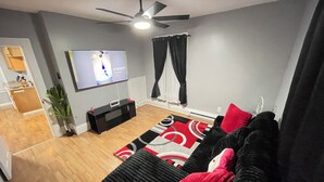 A living room with a black sofa, a red and black patterned rug, a ceiling fan, and a flat-screen TV mounted on the wall.