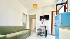Un ambiente compatto con un divano verde, un tavolino con due sedie, una TV fissata al muro e un frigorifero blu.