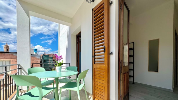 Un balcone con tavolo e sedie, una porta in legno e vista su edifici sullo sfondo.