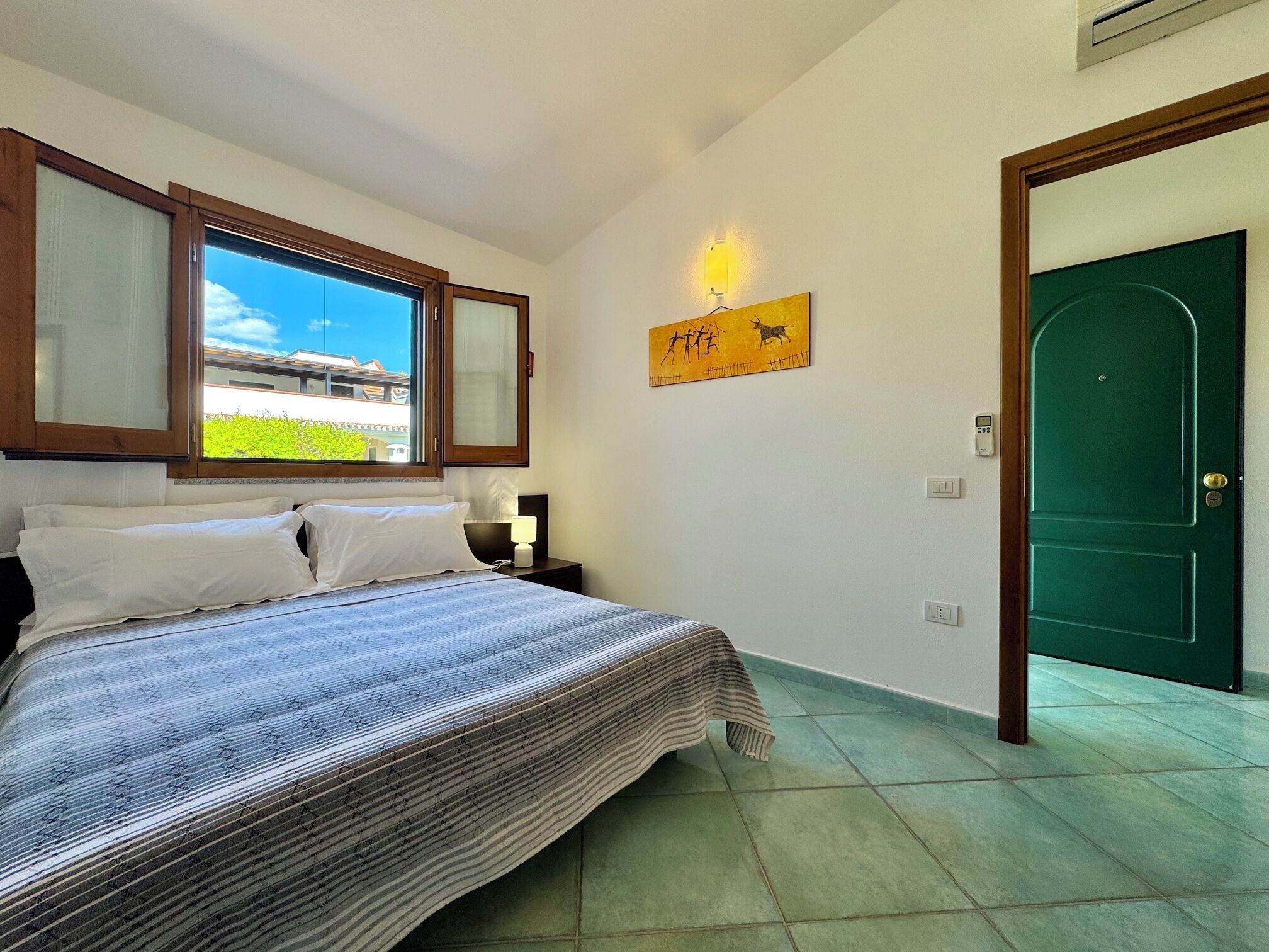 A bedroom with a bed, a window, a green door, and a wall-mounted light fixture.