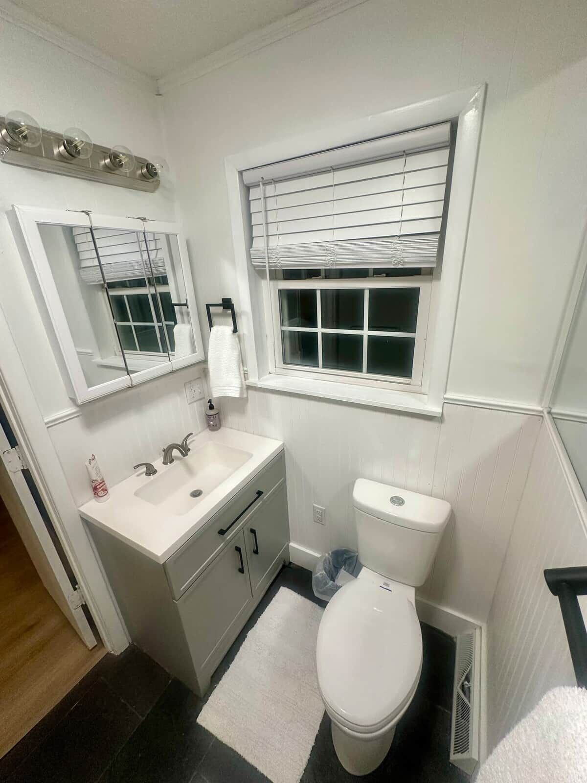 A bathroom with a white sink, mirror, and toilet, a window with blinds, and a towel rack.